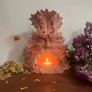 Puede incluir: Un portavelas de cerámica rosa con forma de Hombre Verde con una vela encendida en su interior. El portavelas está decorado con hojas y tiene una cara detallada.