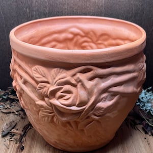 Op de afbeelding: Terracotta bloempot met een reliëf van rozen en bladeren. De pot is lichtbruin van kleur en heeft een glad, licht getextureerd oppervlak.