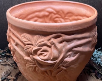 May include: Terracotta flower pot with a raised design of roses and leaves. The pot is a light brown color and has a smooth, slightly textured surface.