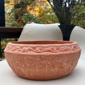 May include: A terracotta planter with a decorative, embossed design of leaves and vines. The planter is round and has a wide, shallow base.
