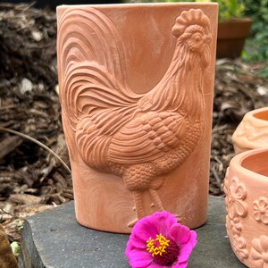 May include: A cylindrical terracotta planter featuring a raised relief design of a rooster on its side. The unglazed clay surface has a natural reddish-brown tone. A single bright pink zinnia flower with a yellow center sits in front.