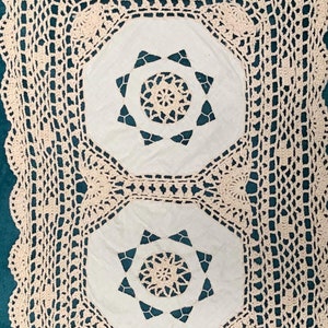 May include: Cream-coloured, handmade crochet table runner. The runner features a central design with two white octagonal panels, each with a starburst pattern. The edges are scalloped, adding a decorative touch. The openwork design is visible throughout.
