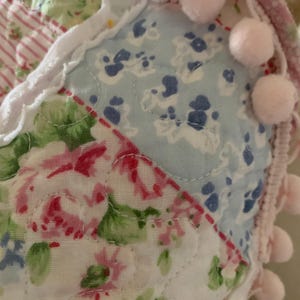 May include: Close-up of a patchwork textile with floral patterns in pink, blue, and green. The edges are trimmed with pink pom-poms and a white lace trim. The fabric is quilted with visible stitching.
