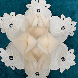 May include: A white, folded, fabric napkin with a delicate floral lace design. The napkin is folded into a square shape with the lace design on all four sides.