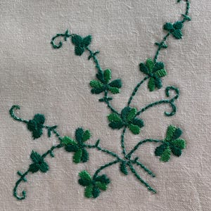 May include: White linen fabric with green embroidered shamrocks and vines. The shamrocks are in a cluster of five, with the vines connecting them.
