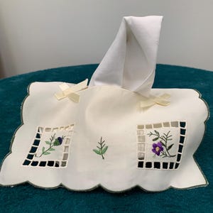 May include: White embroidered tissue box cover with green trim and floral designs. The cover has a scalloped edge and two white satin bows.