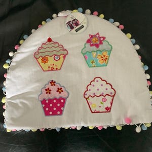 May include: White tea cosy featuring four embroidered cupcake designs in different colours. The cosy is trimmed with colourful pom-poms and has a tag with owl illustrations and the word "COCOON".
