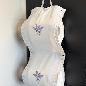 May include: A white fabric toilet paper holder with a lavender embroidery design. The holder has a lace trim and a ribbon for hanging. It is designed to hold two rolls of toilet paper.