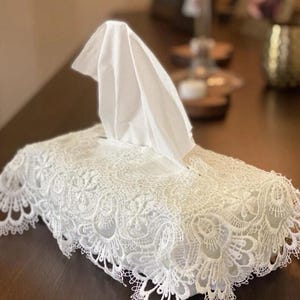 May include: A white lace tissue box cover. The cover is rectangular with a decorative scalloped edge. White tissues are emerging from the top. The lace design is detailed and ornate, adding a touch of elegance.