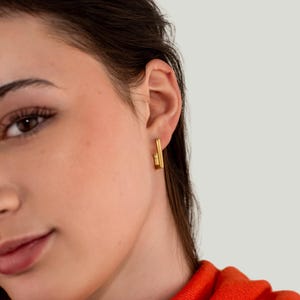May include: A close-up of a person wearing gold earrings. The earrings are rectangular with a vertical bar design. The person is wearing an orange top. The background is a plain, light grey colour.