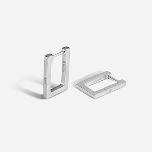 Può includere: Due orecchini a cerchio rettangolari color argento. Ogni orecchino ha una chiusura a cerniera e il testo "925 RO" è visibile sul lato. Gli orecchini sono presentati su sfondo bianco.