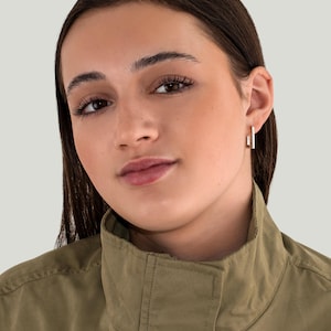 May include: A close-up shot of a person wearing gold-colored, rectangular hoop earrings. The earrings are small and minimalist in design. The person is wearing an olive green jacket.