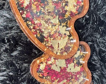 Puede incluir: Juego de bandejas de resina en forma de corazón con borde marrón, relleno de pétalos rojos y copos dorados. Las bandejas están apiladas sobre una superficie negra y difusa. Un artículo decorativo.