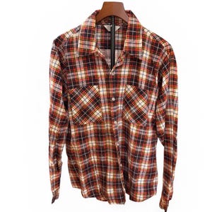 May include: A long-sleeved plaid shirt in a classic pattern of red, black, white, and yellow. The shirt has two chest pockets and a button-down front. The shirt is hanging on a wooden hanger.