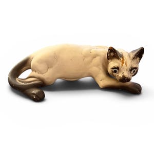 May include: A Siamese cat figurine in a crouching position with a cream-colored body and dark brown points on the ears, face, and tail. The surface shows signs of wear with chipped paint on the ears and face, and the cat has painted eyes.