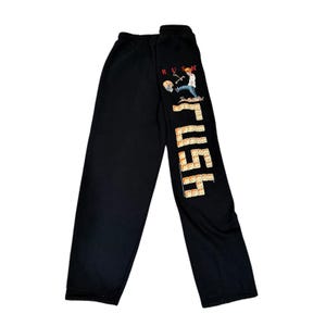 May include: Black sweatpants with the word "RUSH" in a vertical, block-style font. The design includes a cartoon figure kicking a skull and a domino pattern. The text "RUSH" is in red and the dominoes are tan.