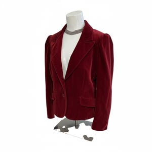 May include: A burgundy velvet blazer with a notched lapel, single-button closure, and two front pockets. The jacket has padded shoulders and is displayed on a white mannequin. A silver necklace is visible.