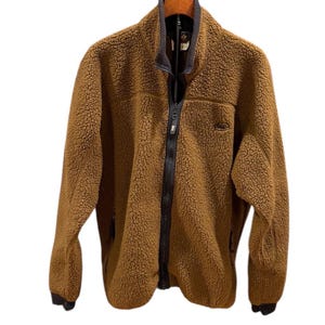 May include: Brown fleece jacket with a full-length black zipper and a small logo on the chest. The jacket has a high collar with a dark gray trim and black cuffs. The jacket is hanging on a wooden hanger.