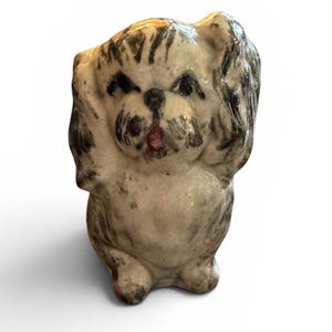 May include: A small ceramic figurine of a dog with a glossy off-white finish and dark gray painted markings. The dog is depicted in a standing position with its front paws raised near its head, featuring a small red painted mouth and dark eyes.