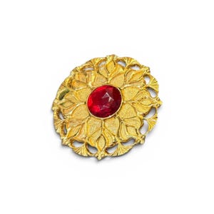 May include: A gold-toned brooch with a central, faceted red gemstone. The brooch features an ornate, floral design with detailed leaf patterns. The overall shape is oval, and the brooch is set against a white background.