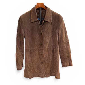 May include: A brown suede jacket with a collared neckline and button-down closure. The jacket is hanging on a wooden hanger against a white background. The jacket has long sleeves and a tailored fit.