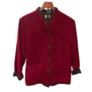 May include: A red velvet jacket with a mandarin collar, long sleeves, and button-down closure. The jacket has two front pockets and dark brown trim along the edges and cuffs. The jacket is hanging on a wooden hanger.