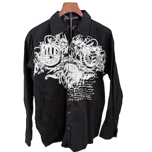 May include: Black button-down shirt with long sleeves and a collared neckline. The shirt features a large white graphic design on the front, with intricate details and script. The shirt is hanging on a wooden hanger.