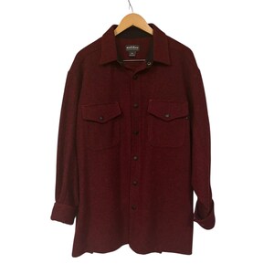 May include: A burgundy long-sleeve shirt with two chest pockets and black buttons. The shirt is made of a textured fabric and features a collar. The brand name "Woolrich" is visible on the label.