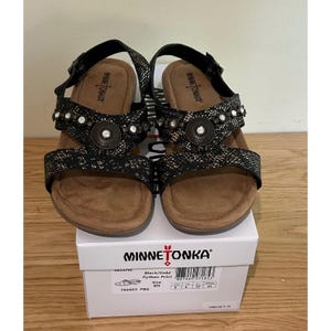 May include: A pair of black and gold Minnetonka Selene sandals with a python print design. The sandals have a buckle closure and decorative embellishments. The sandals are displayed on top of a white box with the Minnetonka logo.