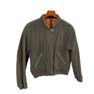 May include: An olive green corduroy bomber jacket featuring a stand-up collar, a front snap button closure, and two slanted zippered pockets. The jacket has an elasticized hem and cuffs, with a contrasting orange interior lining visible at the neck.