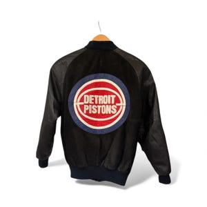 May include: Black bomber jacket with leather sleeves and a ribbed hem. The back features a large Detroit Pistons logo in red, white, and blue. The jacket is hanging on a wooden hanger.