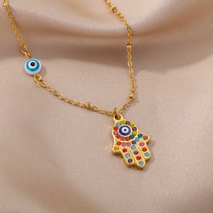 May include: Gold chain necklace with a hamsa hand charm and a blue evil eye charm. The hamsa hand is decorated with colorful rhinestones.