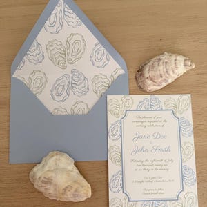 May include: A light blue wedding invitation suite with oyster shell designs. The invitation includes the names Jane Doe and John Smith, with the date and time of the wedding. Two seashells and an envelope are also present.