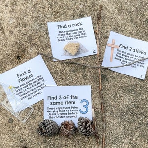 May include: Three white cards with black text on a brown, sandy surface. The cards are part of an Easter scavenger hunt. The cards read: "Find a flower", "Find a rock", and "Find 3 of the same item".