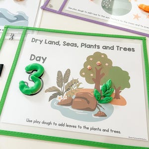 May include: A green felt board with a white background and the text "Dry Land, Seas, Plants and Trees" and "Day 3". The board has a green border and a clear plastic overlay. There are images of a tree, a pond, and rocks. The text "Use play dough to add leaves to the plants and trees." is at the bottom of the board.