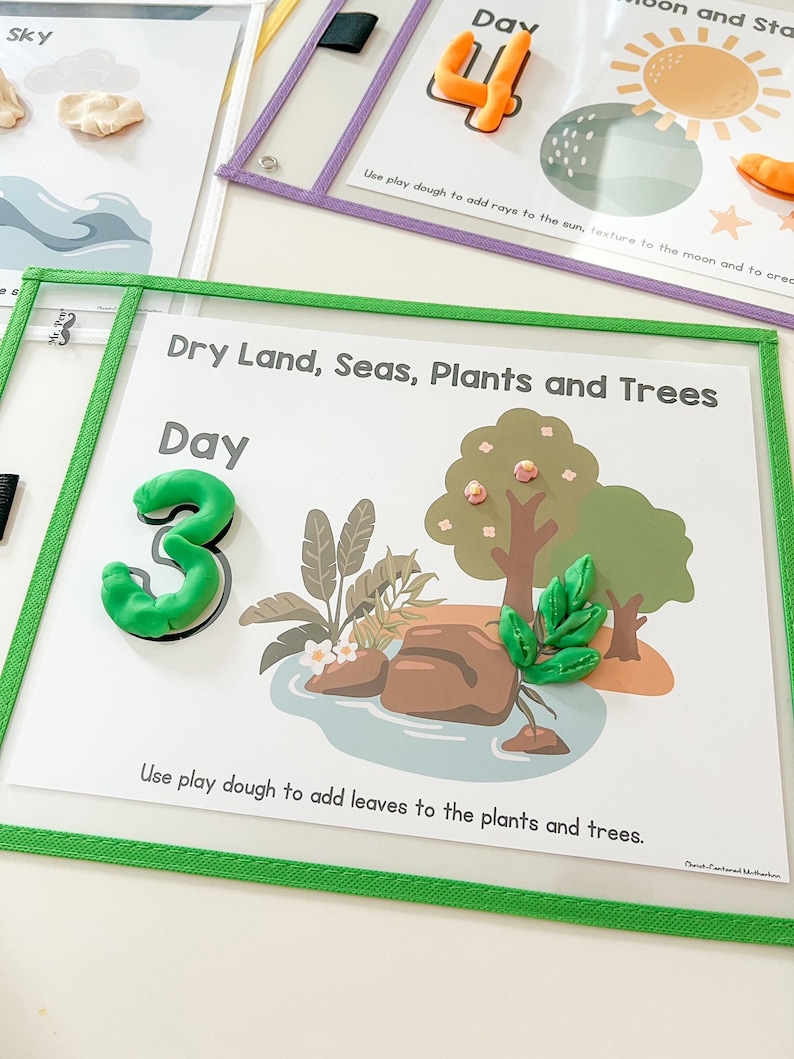 May include: A green felt board with a white background and the text "Dry Land, Seas, Plants and Trees" and "Day 3". The board has a green border and a clear plastic overlay. There are images of a tree, a pond, and rocks. The text "Use play dough to add leaves to the plants and trees." is at the bottom of the board.