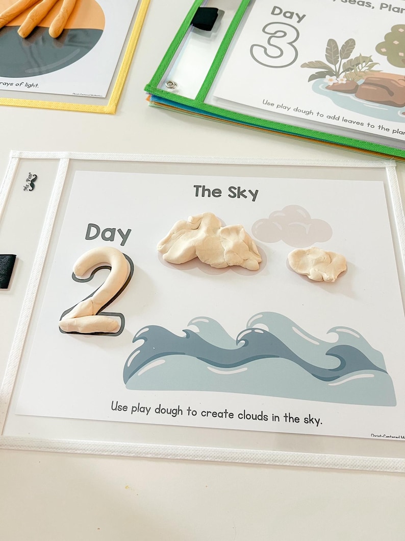 May include: A white plastic frame with a clear plastic sheet inside. The sheet has a blue and white illustration of waves and the text "The Sky". There is a white play dough number "2" and the text "Day 2" on the left side of the sheet. There is a white play dough cloud on the right side of the sheet. The text "Use play dough to create clouds in the sky." is at the bottom of the sheet.