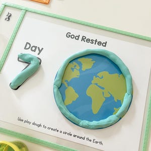 May include: A white card with the text "Day 7" and "God Rested" written on it. A blue and green globe is surrounded by a circle of green play dough. The text "Use play dough to create a circle around the Earth." is written below the globe.