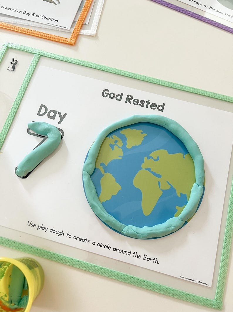 May include: A white card with the text "Day 7" and "God Rested" written on it. A blue and green globe is surrounded by a circle of green play dough. The text "Use play dough to create a circle around the Earth." is written below the globe.