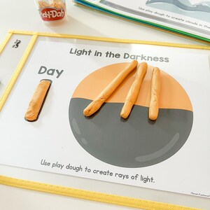 May include: A white card with the text "Light in the Darkness" and "Day" printed on it. There is a grey circle with orange play dough rays of light coming out of it. The card is on a yellow surface.