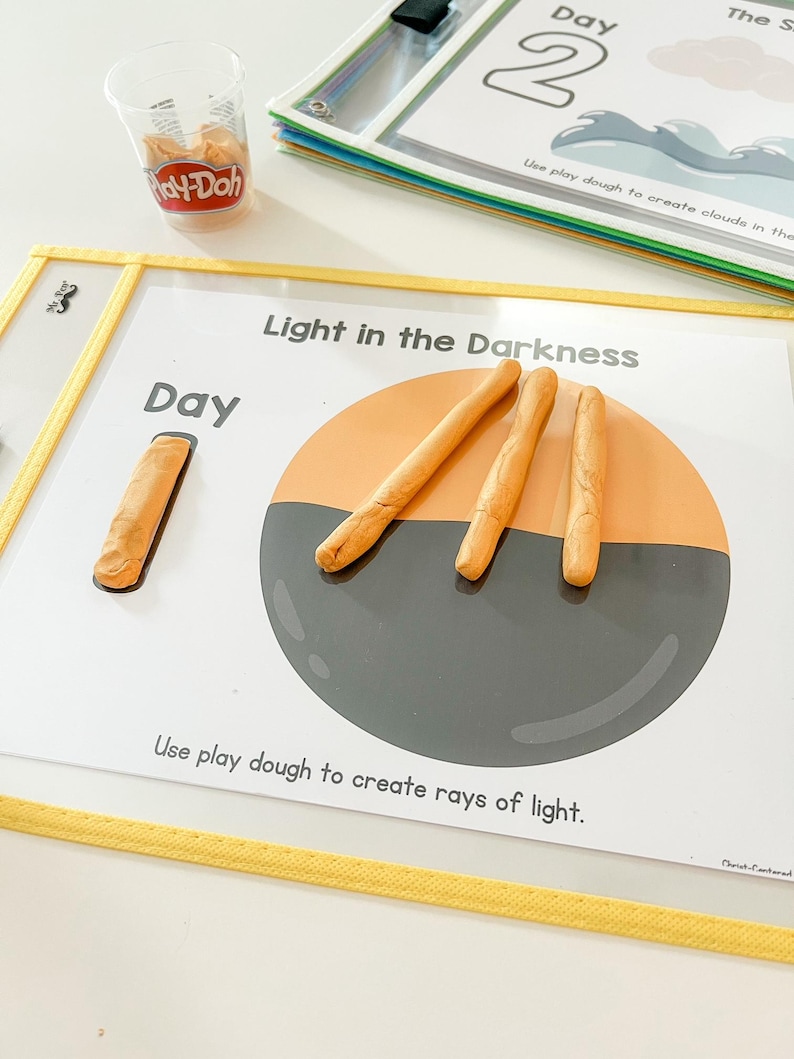 May include: A white card with the text "Light in the Darkness" and "Day" printed on it. There is a grey circle with orange play dough rays of light coming out of it. The card is on a yellow surface.