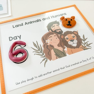 May include: A white card with the text "Land Animals and Humans" and "Day 6" in black. The card has a brown bear, a lion, and a man and woman with brown hair. The card has a pink play dough number 6 and instructions to add another animal using play dough.
