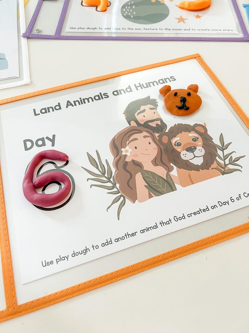 May include: A white card with the text "Land Animals and Humans" and "Day 6" in black. The card has a brown bear, a lion, and a man and woman with brown hair. The card has a pink play dough number 6 and instructions to add another animal using play dough.