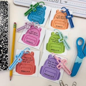 May include: A collection of six square cards featuring colorful backpack illustrations with religious text and scripture references, each attached to a matching plastic cross keychain. The cards include phrases like Let your light shine and Put on the full armor of God.