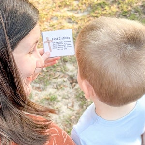 May include: A woman and a young boy are playing a game outdoors. The woman is holding a card that says "Find 2 sticks. Use the sticks to make the shape of a cross. These represent when Jesus died on the cross for our sins."