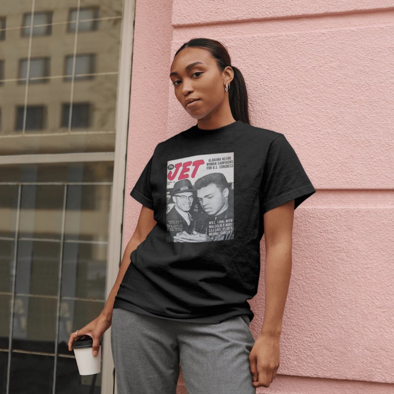 May include: A black t-shirt with a vintage Jet magazine cover featuring a photo of Muhammad Ali and Malcolm X. The magazine cover text reads "Alabama News,  W.L.  Norton  Running  For U.S. Congress,  Will Link With Malcolm X,  Celebrates  King's  Birthday".