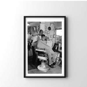 May include: Black and white photo of a barber cutting a young man's hair in a barbershop. The barber is standing and the young man is sitting in a barber chair.