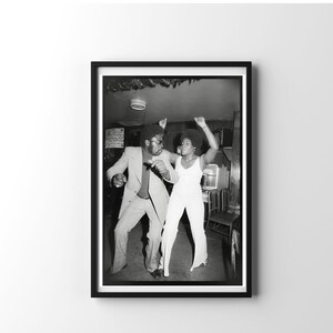 May include: A black and white photograph of a man and a woman dancing in a dimly lit room. The man is wearing a suit and the woman is wearing a white jumpsuit. They are both smiling and having a good time.