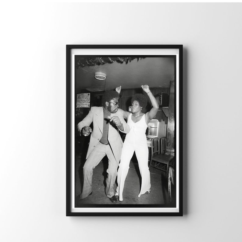 May include: A black and white photograph of a man and a woman dancing in a dimly lit room. The man is wearing a suit and the woman is wearing a white jumpsuit. They are both smiling and having a good time.