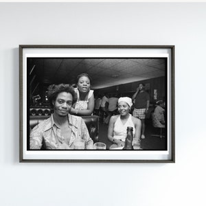 May include: Black and white photo of three people sitting at a bar. The person in the foreground is wearing a patterned shirt and looking at the camera. The person behind them is wearing a white shirt and looking at the camera. The person in the background is wearing a white hat and looking at the camera.